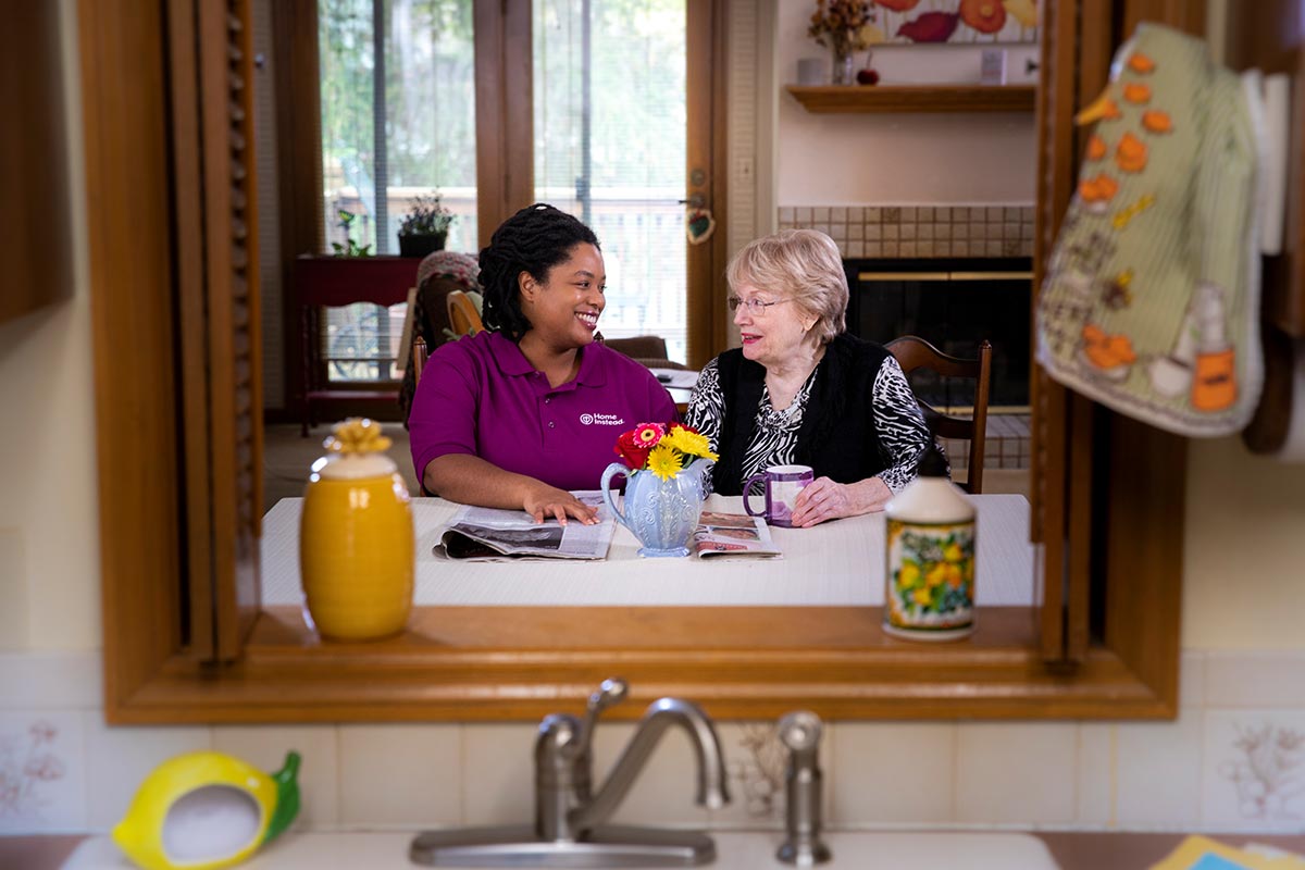 Home Instead caregiver and senior woman sit talking at kitchen table